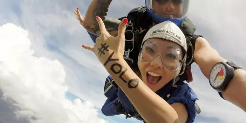 Excited woman in tandem skydive with #YOLO written on arm during freefall over Auckland