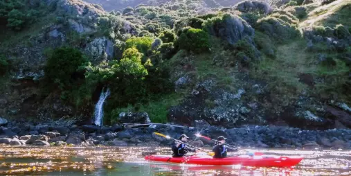 Two people kayaking past a small waterfall surrounded by lush cliffs in Akaroa