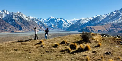 Two people pose with swords on a grassy ridgeline with snow-capped mountains behind.