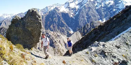 Two hikers making their way up the rocky trail on Mount Sebastopol with snow-covered peaks in the background.