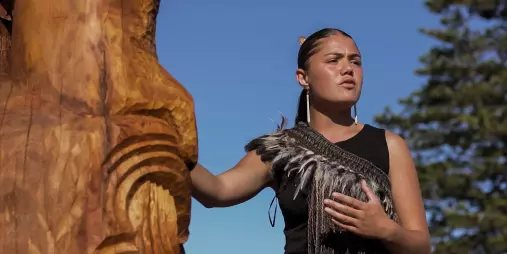 Māori guide delivering a cultural welcome beside a carved wooden figure during a Manea tour in Northland