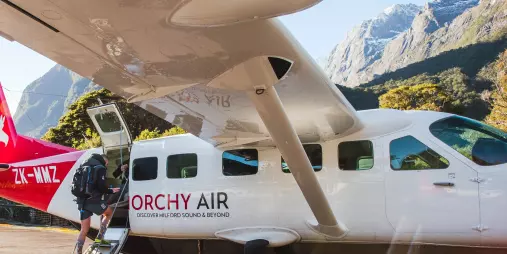 Passenger boarding a Glenorchy Air flight at Milford Sound airstrip with scenic peaks in the background.