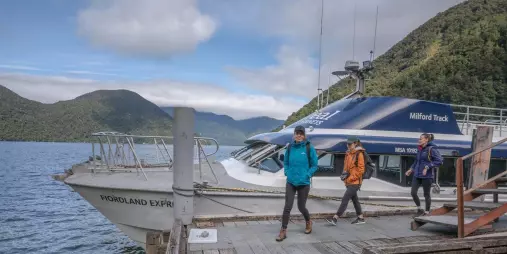 Three hikers walking toward the Fiordland Express, Milford Track Water Taxi, docked at a scenic pier.
