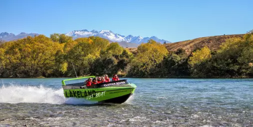Jet boat speeding along the Clutha River with passengers on board and snow-capped mountains in the background.