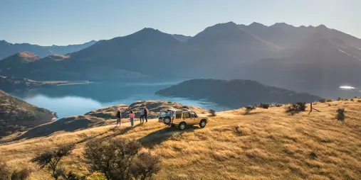 A 4WD vehicle parked on a grassy hilltop with people enjoying sweeping views over Lake Wanaka.