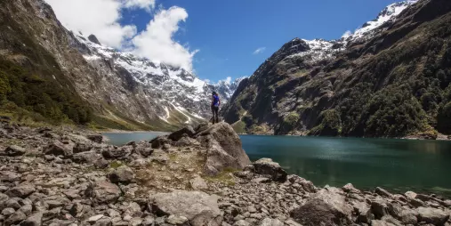 Walker stands on rocky shoreline above Lake Marian surrounded by snow-capped mountains.