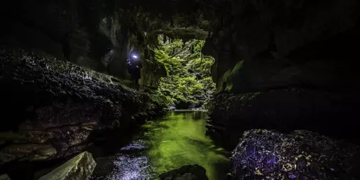 A photographer with a headlamp captures images inside a Waitomo limestone cave, featuring a glowing green stream and moss-covered walls.