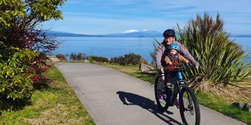Parent and child cycling along a paved trail by Lake Taupō, with snow-capped mountains visible in the distance.