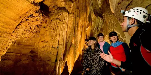 Waitomo guide explaining cave formations to a group of visitors wearing helmets inside an illuminated limestone cave